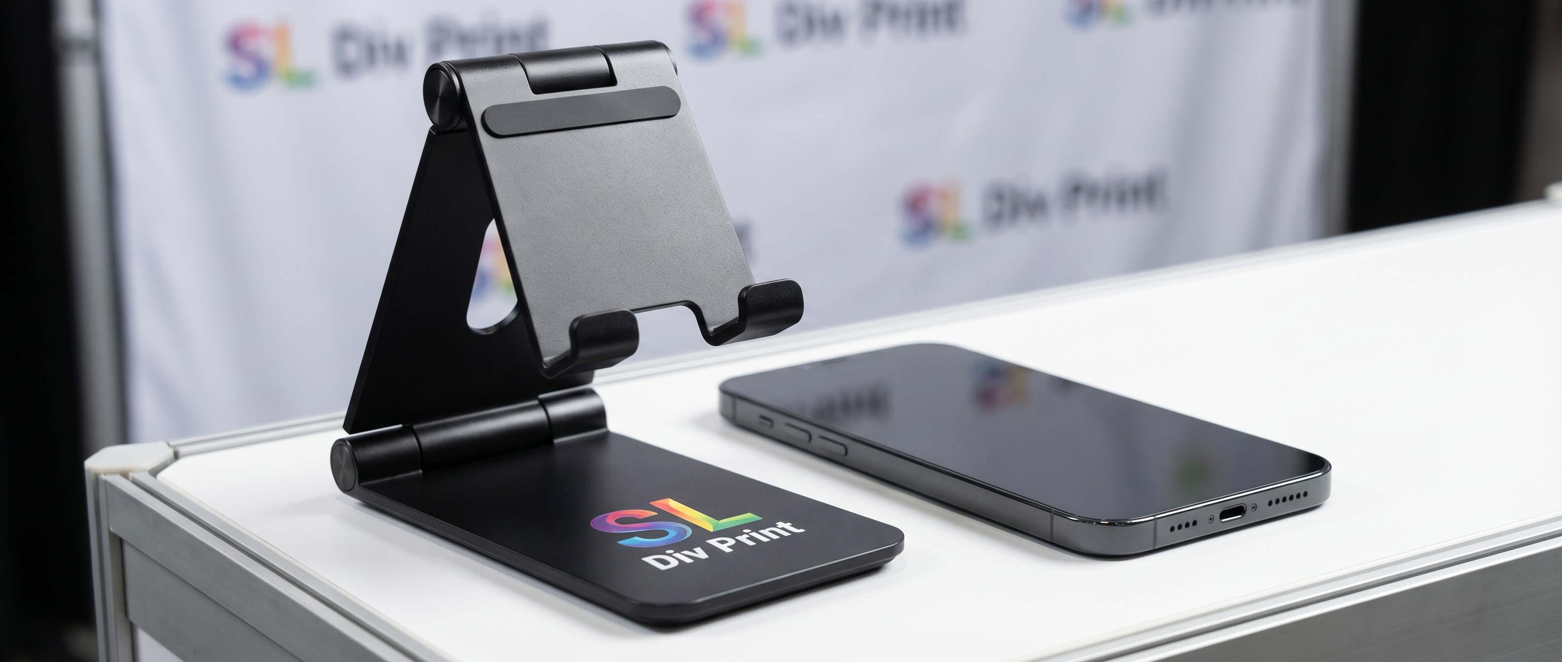 Adjustable black phone stand on a white desk with a glossy smartphone beside it and a DIY Print base logo nearby trade show booth promotional products portable phone stand 2026 trade show giveaways 2026 promotional products for trade shows best trade show swag trade show booth promotional products top trade show giveaways effective trade show promotional products rush promotional products for trade shows sustainable trade show promotional products trade show swag ideas 2026 best promotional giveaways for trade shows