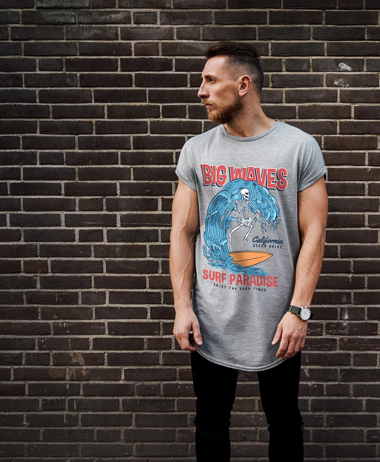 Man with short hair and beard standing against a brick wall wearing a gray graphic Tshirt that reads BIG WAVES and shows a surfing skeleton image beside the words Surf Paradise