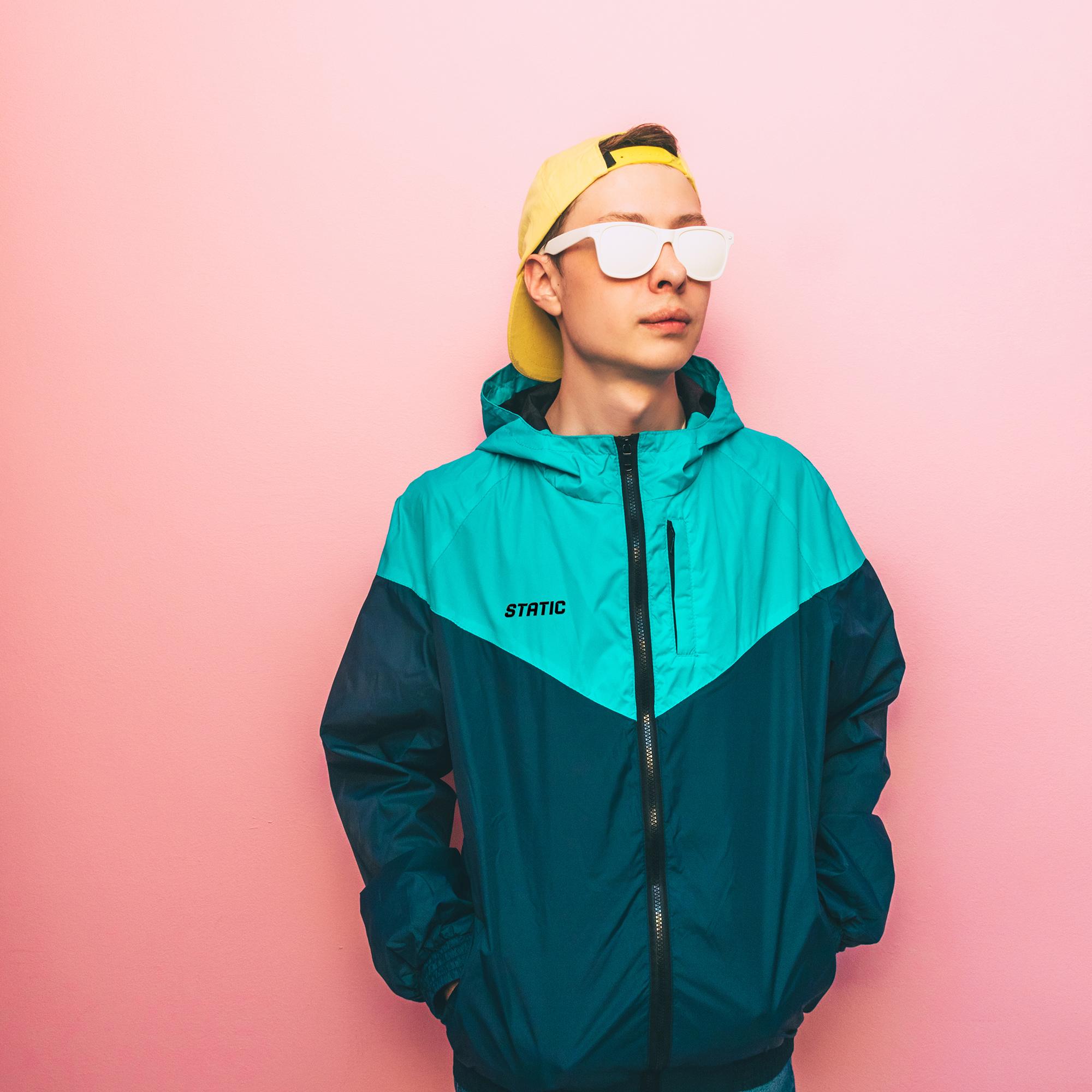 Young person wearing a turquoise and navy color block windbreaker yellow backward cap and white sunglasses against a pink wall looking to the right