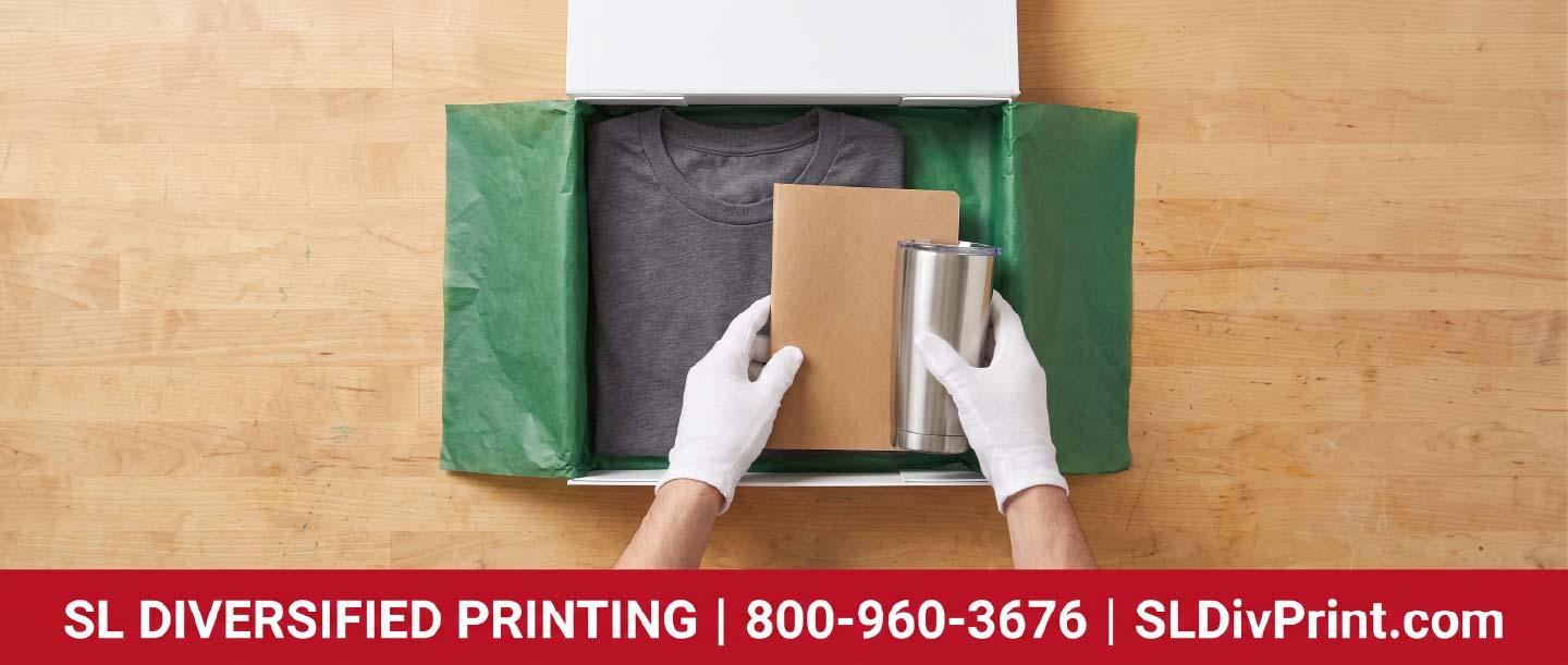 Gloved hands arranging folded apparel a notebook and tissue paper inside an open branded gift box on a workbench employee swag fulfillment remote team merch branded merchandise fulfillment merch warehousing company store kitting Arizona distribution hub onboarding kit shipping international merch delivery promotional product storage