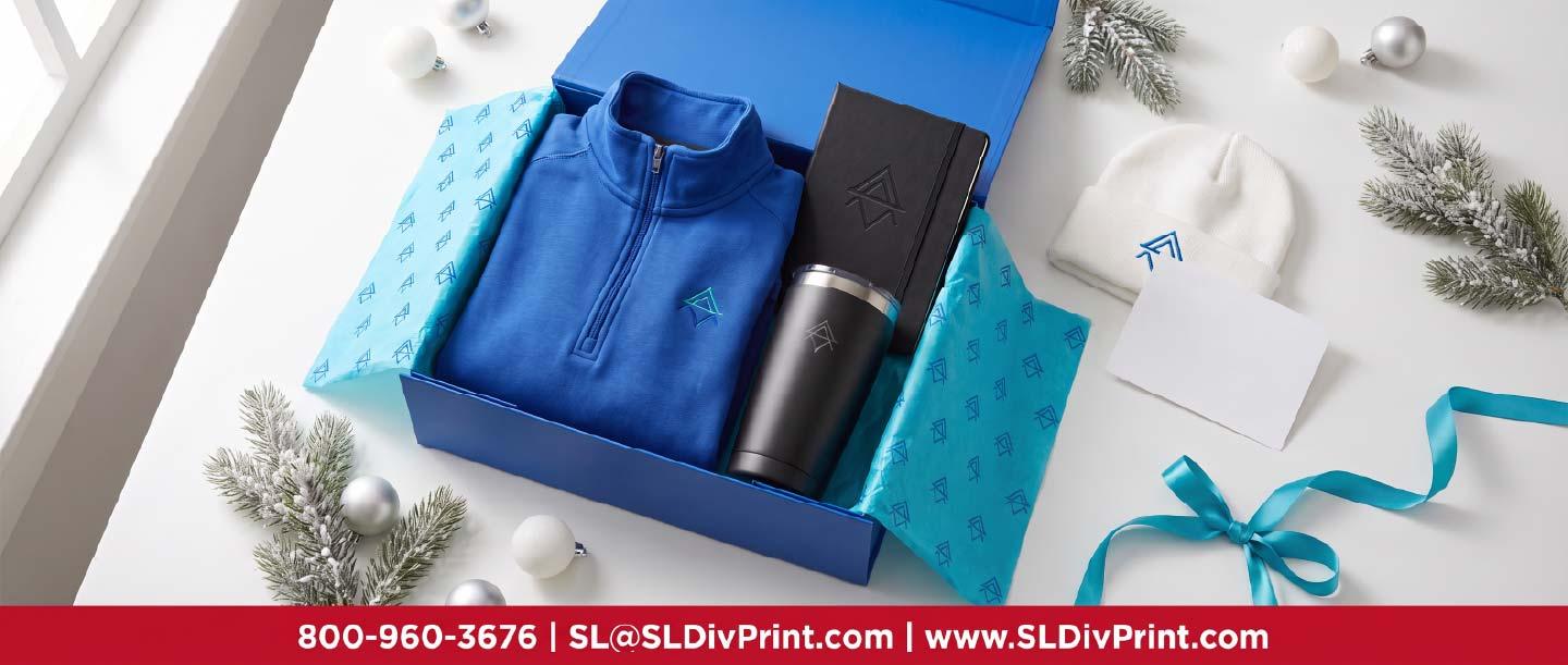 Corporate Holiday Gifts Branded Gift Box | SL Diversified Printing Open royal blue gift box with an embroidered quarter zip black tumbler notebook knit beanie and card on a white table corporate holiday gifts employee appreciation client retention repeat business branded swag custom apparel promotional products rush printing order fulfillment logo merchandise remote teams
