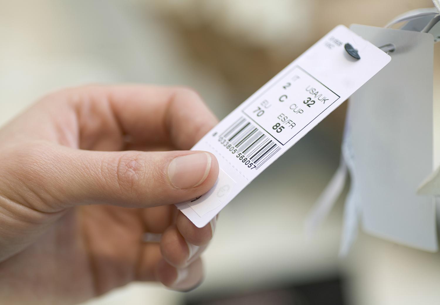 Close up of a hand holding a white tag with a barcode and numbers attached to a product or tag strap