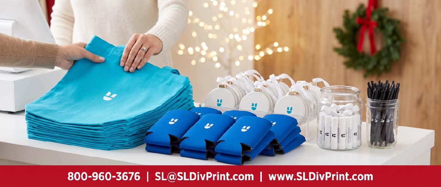 Holiday Promotional Products for Customer Giveaways | SL Diversified Printing Store counter with teal tote bags blue can coolers white ornaments lip balms and pens as a hand reaches for a tote bag corporate holiday gifts employee appreciation client retention repeat business branded swag custom apparel promotional products rush printing order fulfillment logo merchandise remote teams